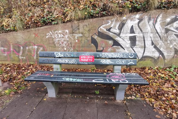 Parkbank, mit rotem Schild auf dem Notfallnummer der Feuerwehr und Standortnummer der Parkbank gedruckt sind, Parkbank ist voll mit Graffiti-Tags
