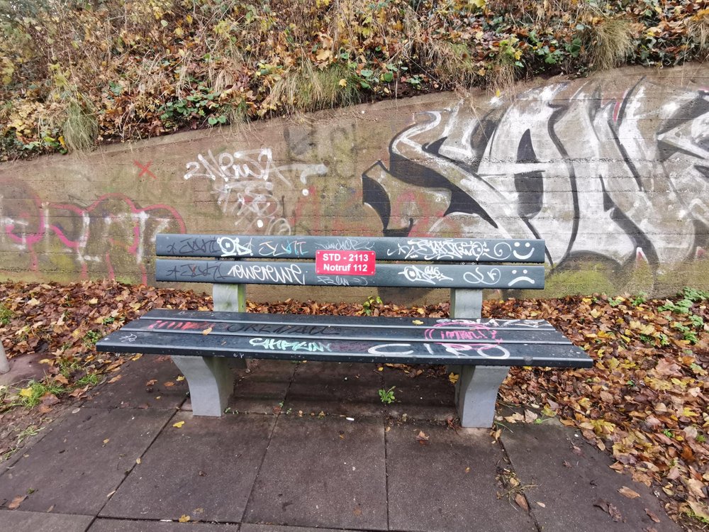Parkbank, mit rotem Schild auf dem Notfallnummer der Feuerwehr und Standortnummer der Parkbank gedruckt sind, Parkbank ist voll mit Graffiti-Tags