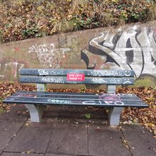 Parkbank, mit rotem Schild auf dem Notfallnummer der Feuerwehr und Standortnummer der Parkbank gedruckt sind, Parkbank ist voll mit Graffiti-Tags